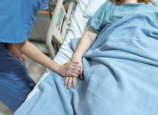 Mercer professors awarded grant to explore how to better support dying patients A health care worker holds the hand of a patient lying in a hospital bed covered with a blue blanket.