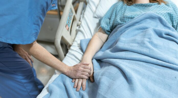 Mercer professors awarded grant to explore how to better support dying patients A health care worker holds the hand of a patient lying in a hospital bed covered with a blue blanket.