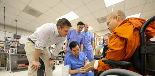 A group of medical professionals shows a prosthetic leg to a person in a wheelchair inside a lab.