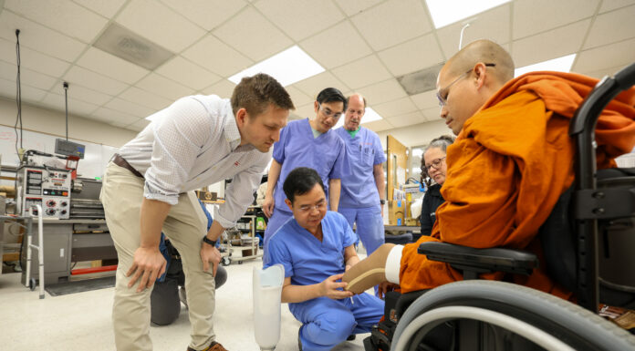 Mercer On Mission team fits monk injured in peace walk with prosthetic leg A group of medical professionals shows a prosthetic leg to a person in a wheelchair inside a lab.