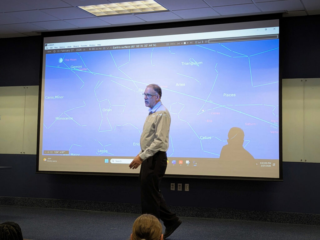 A man stands in front of a large screen displaying a digital star map with constellations and planets labeled.