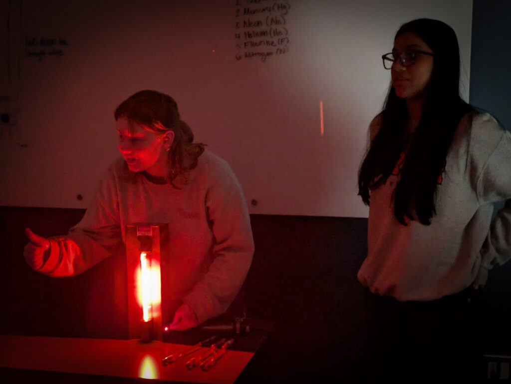 Two people stand near a glowing red light tube in a dark room, with a whiteboard in the background.
