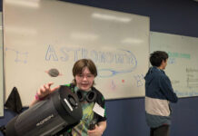 Mercer students, faculty bring space science to Girl Scouts Student holding a telescope in front of a whiteboard with ASTRONOMY and space-themed drawings; another student stands nearby.