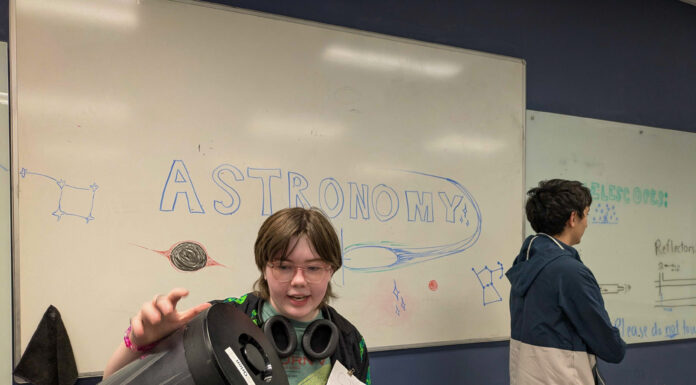 Mercer students, faculty bring space science to Girl Scouts Student holding a telescope in front of a whiteboard with ASTRONOMY and space-themed drawings; another student stands nearby.
