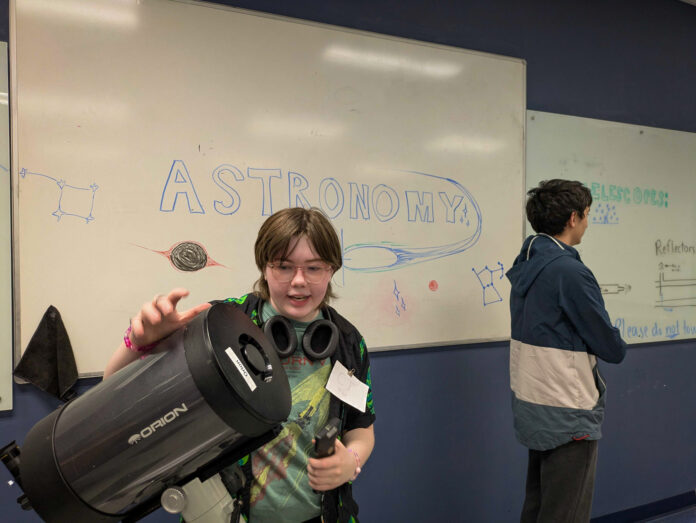 Student holding a telescope in front of a whiteboard with ASTRONOMY and space-themed drawings; another student stands nearby.