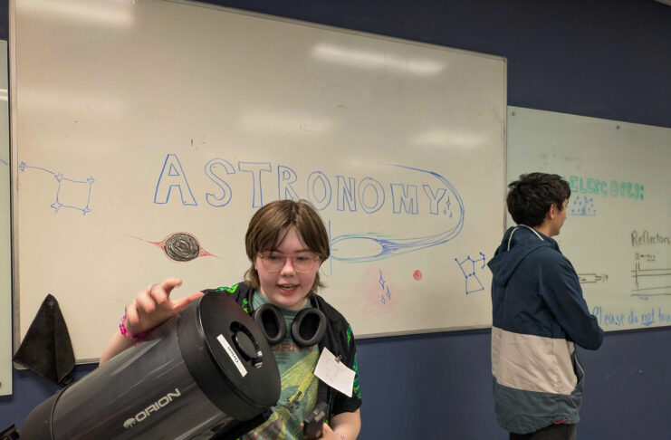 Student holding a telescope in front of a whiteboard with ASTRONOMY and space-themed drawings; another student stands nearby.