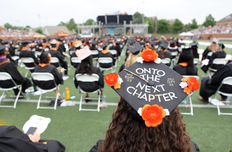 Graduate wearing a decorated cap reading Onto the Next Chapter at an outdoor commencement ceremony with seated attendees.