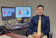 Mercer pharmacy professor named Fellow of the American Heart Association Man in business attire sits at a desk with documents, glasses, and two monitors displaying medical diagrams behind him.