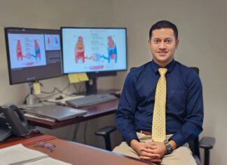 Mercer pharmacy professor named Fellow of the American Heart Association Man in business attire sits at a desk with documents, glasses, and two monitors displaying medical diagrams behind him.