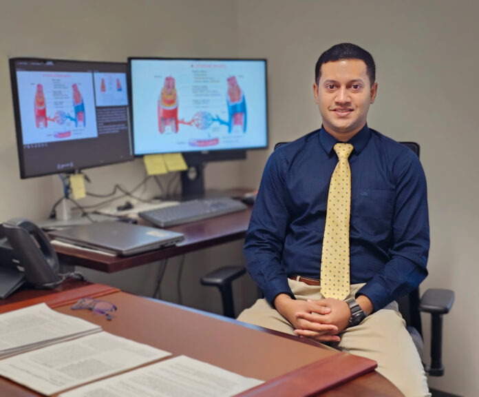 Man in business attire sits at a desk with documents, glasses, and two monitors displaying medical diagrams behind him.