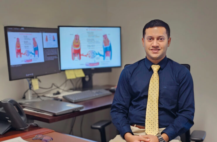 Man in business attire sits at a desk with documents, glasses, and two monitors displaying medical diagrams behind him.