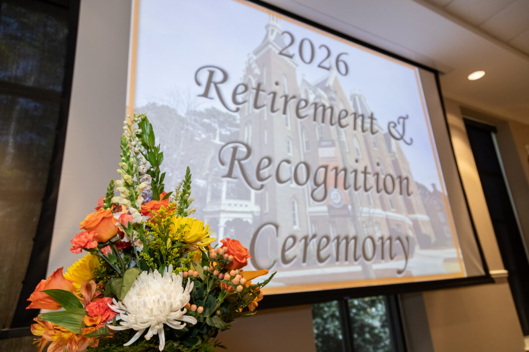 A flower arrangement in front of a screen displaying 2026 Retirement & Recognition Ceremony in a conference room.