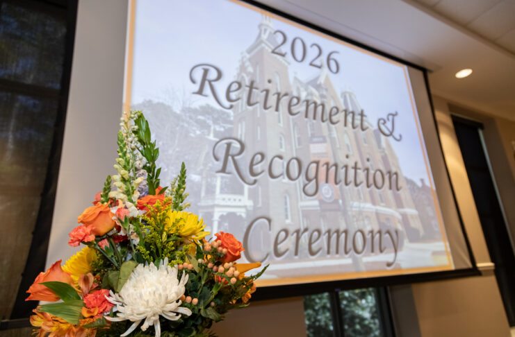 A flower arrangement in front of a screen displaying 2026 Retirement & Recognition Ceremony in a conference room.