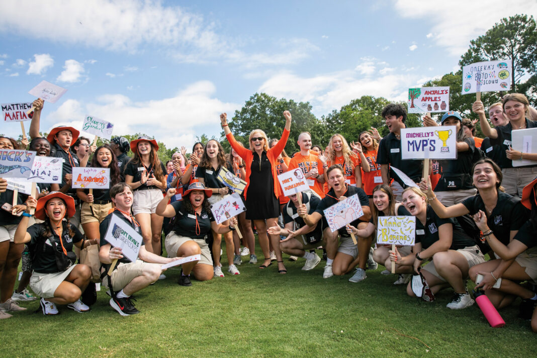 A group of smiling young people, mostly in black shirts and khaki shorts, hold up signs with various names and drawings.