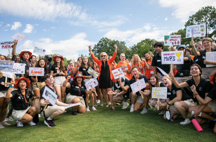 A group of smiling young people, mostly in black shirts and khaki shorts, hold up signs with various names and drawings.