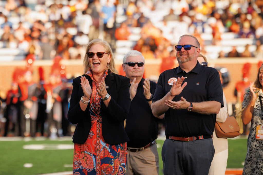 Three adults stand on a football field clapping, with a crowd and marching band blurred in the background.