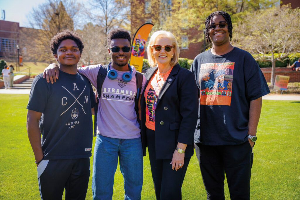Four people stand together outdoors on a sunny day, smiling at the camera with trees and buildings in the background.