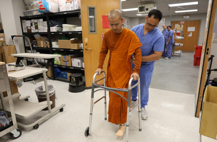 A man wearing scrubs assists a monk with a prosthetic leg as he walks using a walker in a lab.