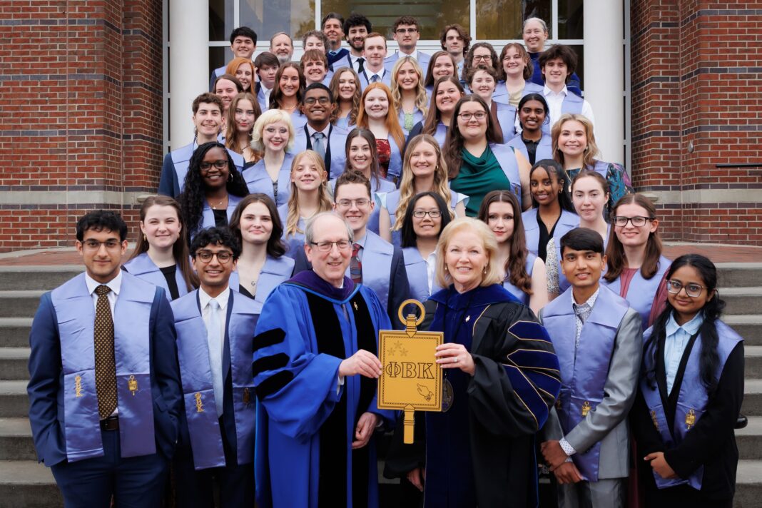 A group of students and faculty in academic regalia pose on steps, holding a large Phi Beta Kappa key symbol.