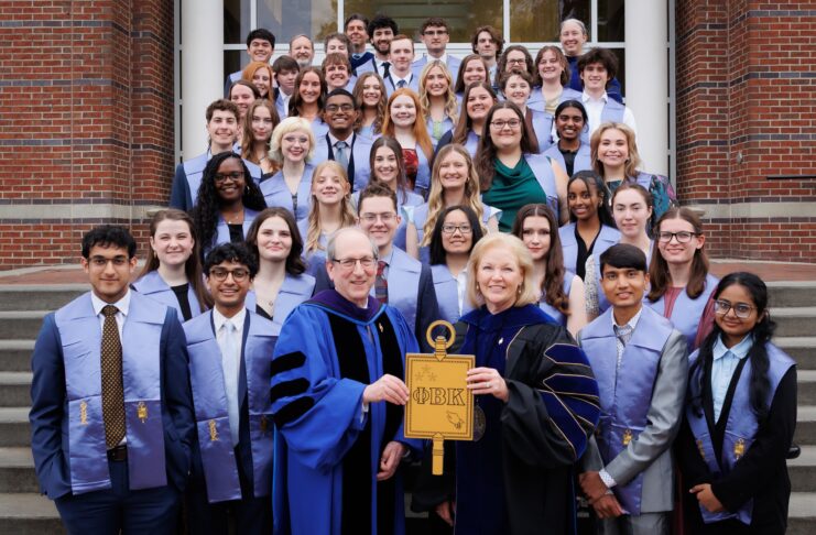 A group of students and faculty in academic regalia pose on steps, holding a large Phi Beta Kappa key symbol.