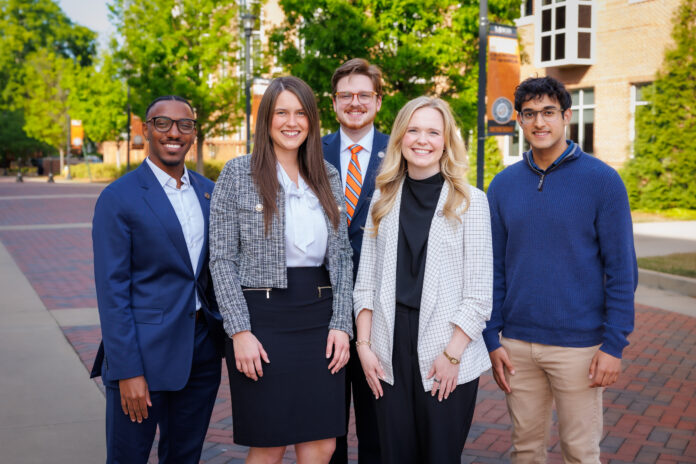 Five professionally dressed people stand and smile outside on a brick walkway with trees and buildings in the background.