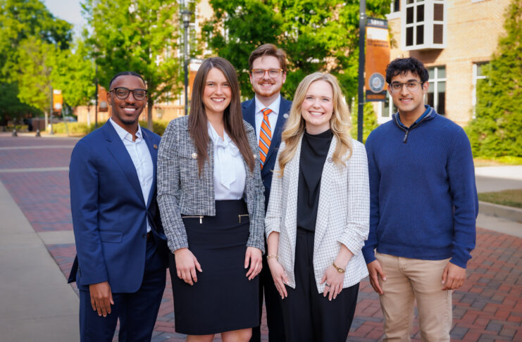 Five professionally dressed people stand and smile outside on a brick walkway with trees and buildings in the background.