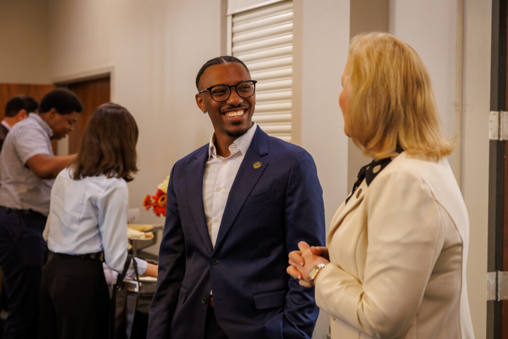 Two people in business attire are talking and smiling at an indoor event while others serve themselves food in the background.