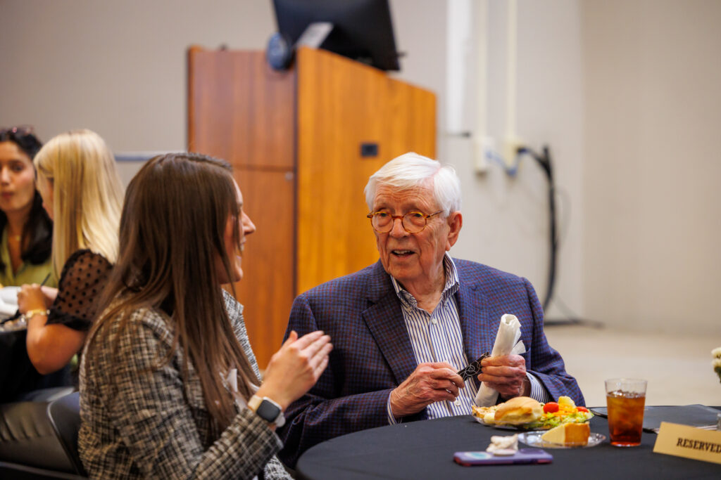 An older man and a woman sit at a table with food and drinks, talking during an indoor event.