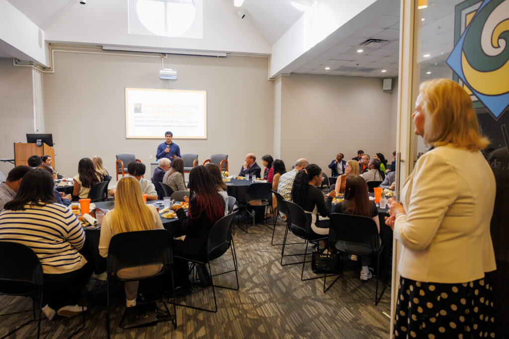 A group of people seated at tables listen to a speaker presenting at the front of a conference room.
