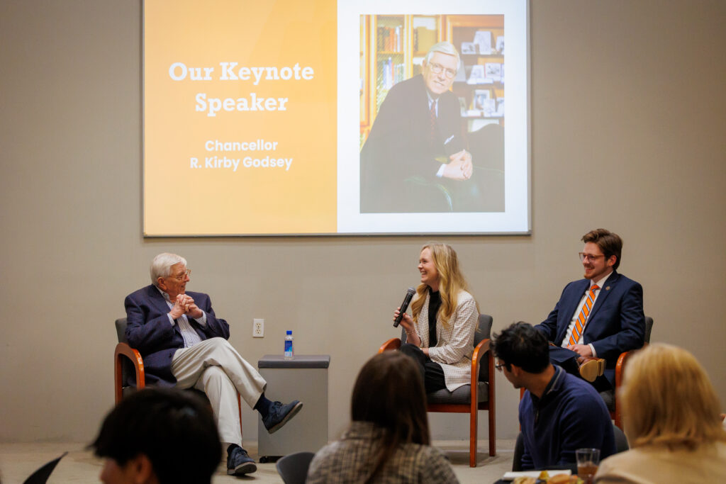 Three people sit on stage in discussion, with a presentation slide showing Our Keynote Speaker: Chancellor R. Kirby Godsey.