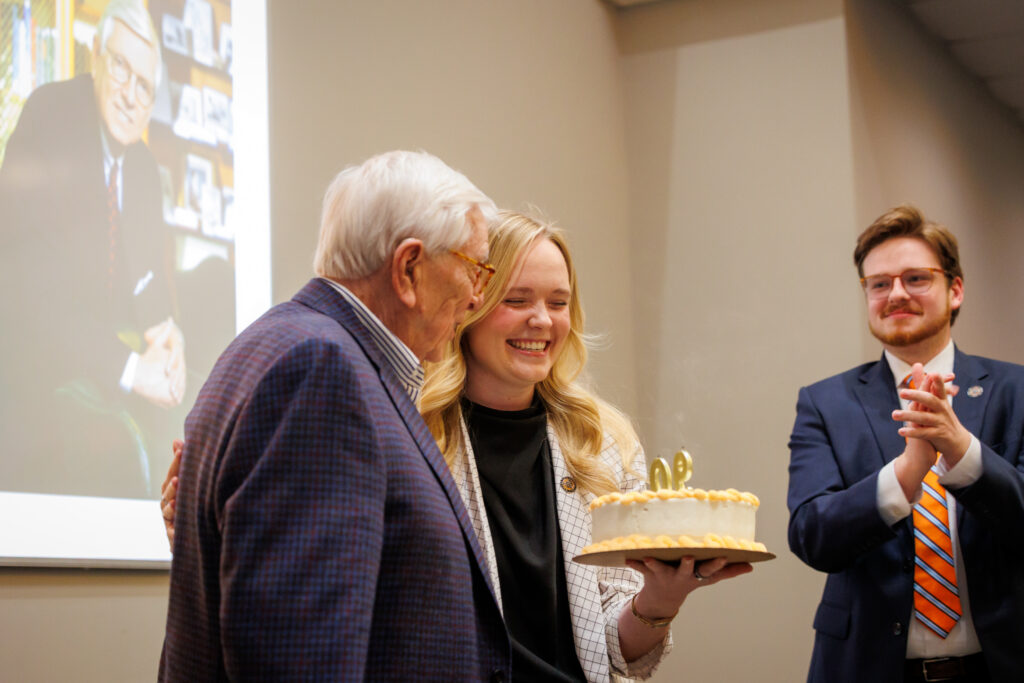 An older man and a woman hold a birthday cake with candles while a man in a suit claps in the background.