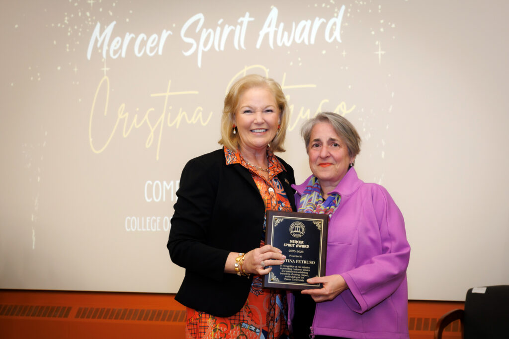 Two women stand together smiling, one holding a Mercer Spirit Award plaque, with a presentation screen in the background.