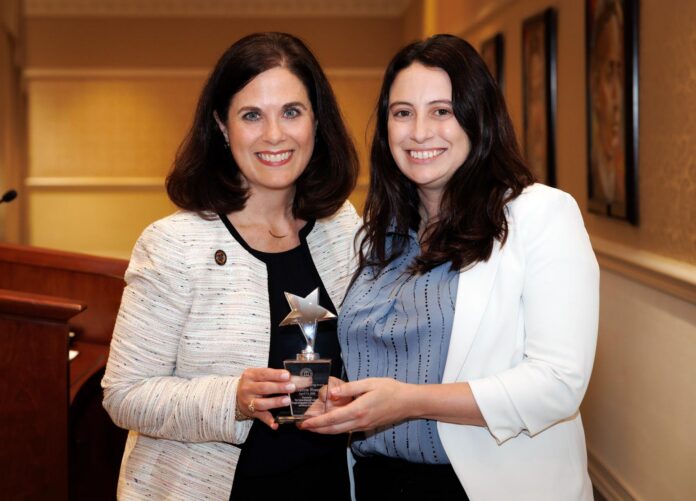 Two women standing indoors, smiling and holding a star-shaped award together in front of a podium.