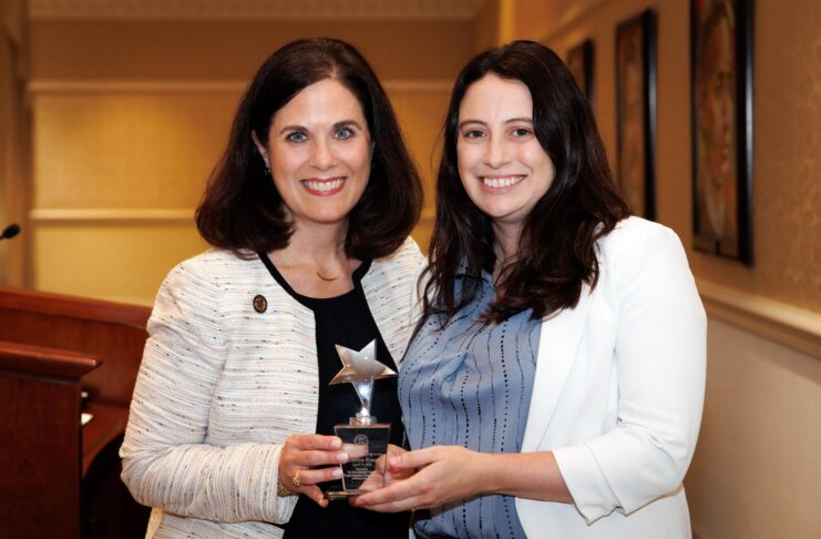 Two women standing indoors, smiling and holding a star-shaped award together in front of a podium.