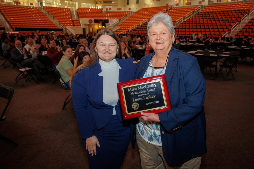 Two women stand in an auditorium; one holds a plaque reading Mike MacCarthy Mentorship Award, Laura Lackey, April 14, 2023.
