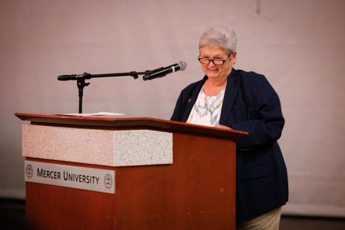An older woman stands at a podium labeled Mercer University, speaking into a microphone.