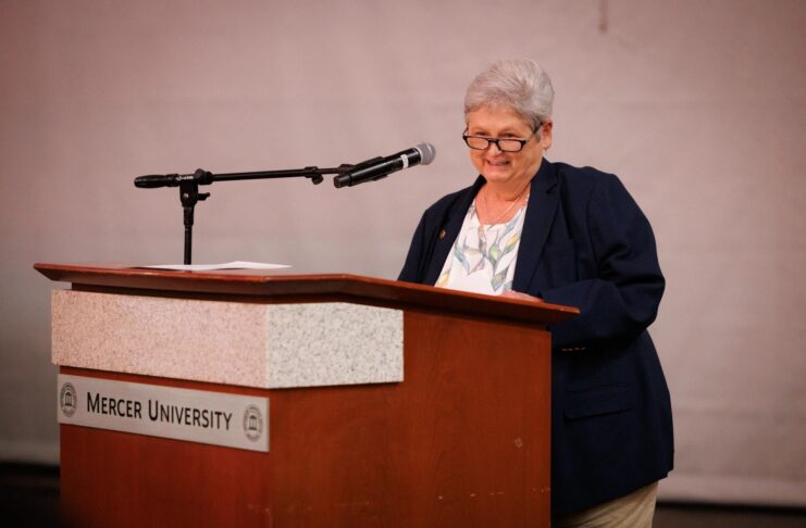 An older woman stands at a podium labeled Mercer University, speaking into a microphone.