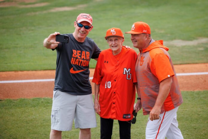 Three people in Mercer University baseball attire pose for a selfie on a baseball field near the foul line.