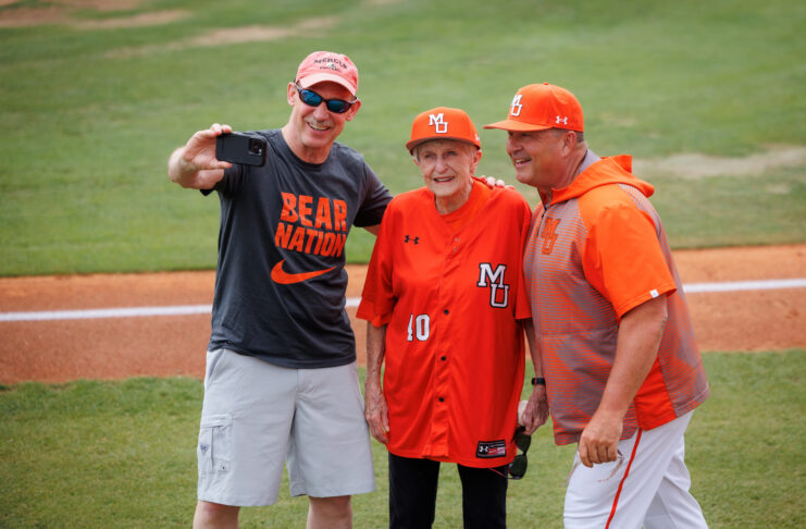 Three people in Mercer University baseball attire pose for a selfie on a baseball field near the foul line.