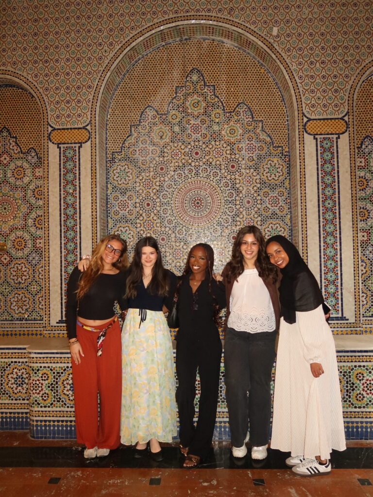 Five women stand side by side in front of a decorative, colorful mosaic tiled wall.