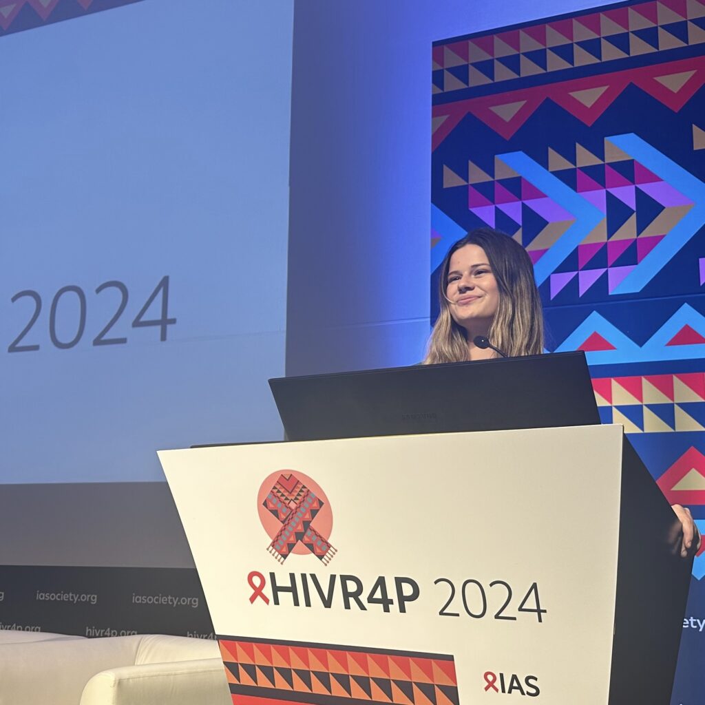 Woman standing at a podium labeled HIVR4P 2024 with conference branding and geometric patterns in the background.
