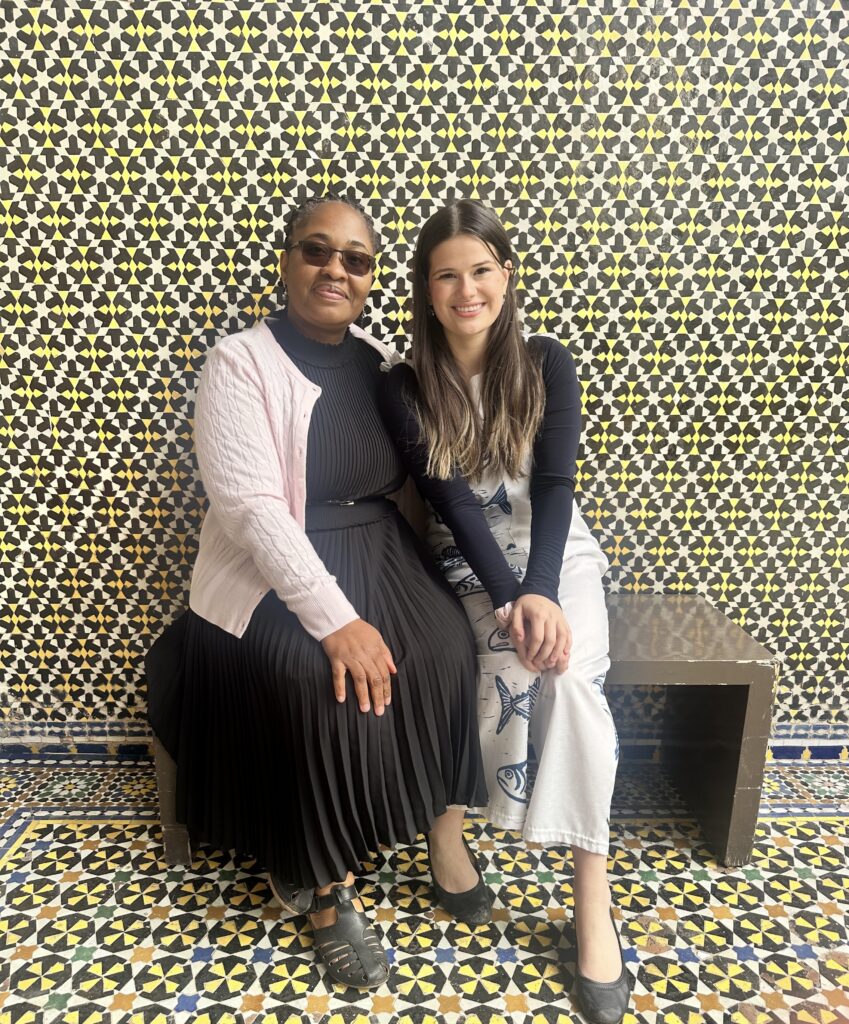 Two women sit side by side on a bench in front of a decorative, geometric tiled wall and floor.
