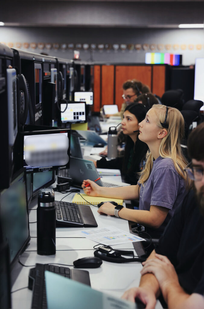 People working at computer stations in an office, focusing on monitors and taking notes.