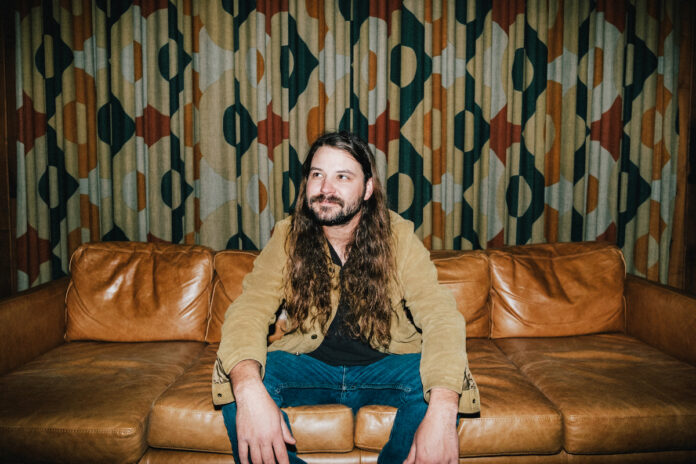 Brent Cobb sits on a leather couch in front of patterned curtains.