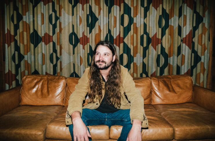 Brent Cobb sits on a leather couch in front of patterned curtains.