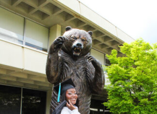 Graduate Spotlight: Brittany Jones Brittany Jones in cap and gown poses in front of a large bear statue on a brick walkway outside a building.