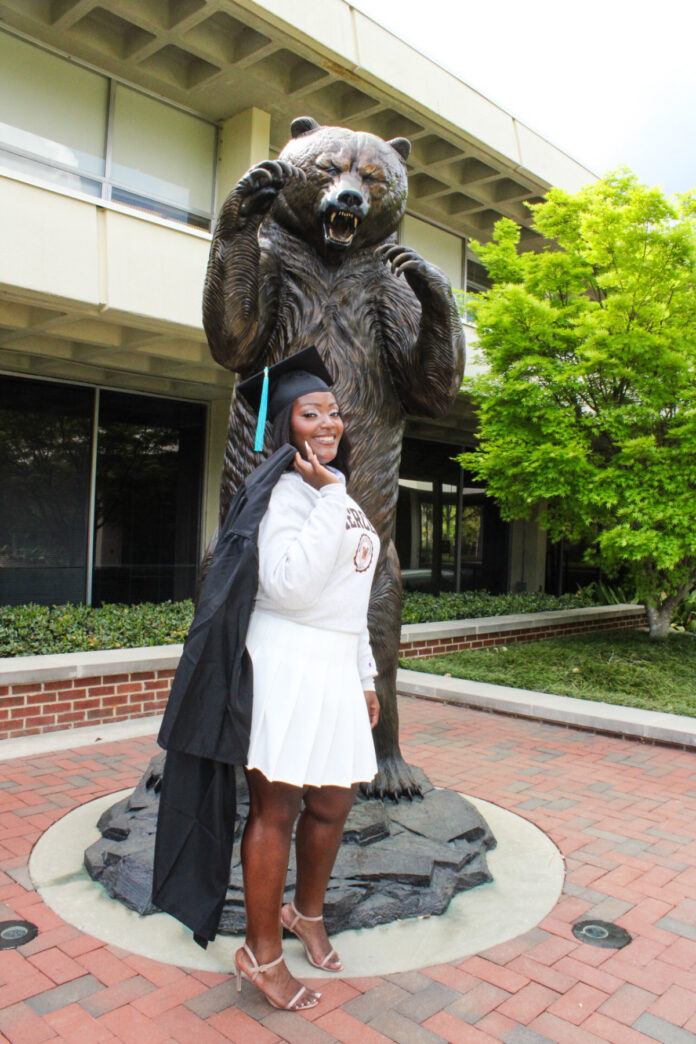 Brittany Jones in cap and gown poses in front of a large bear statue on a brick walkway outside a building.