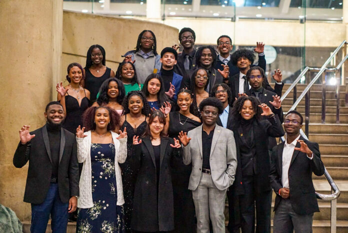 A group of Mercer University students pose on stairs indoors, dressed formally and making a claw gesture with their hands.