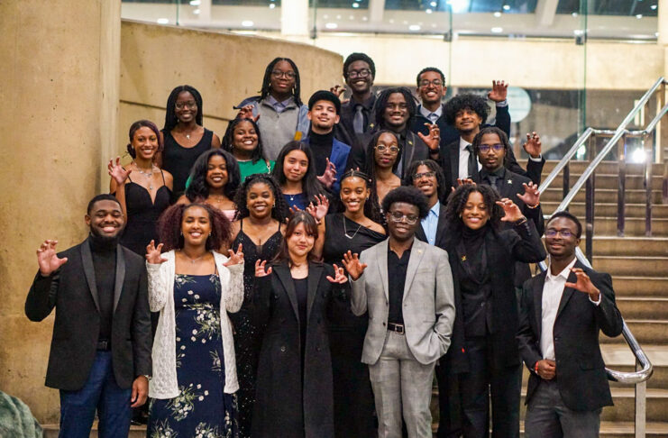 A group of Mercer University students pose on stairs indoors, dressed formally and making a claw gesture with their hands.