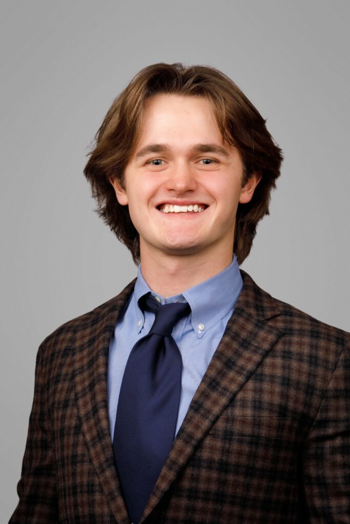 Young man with medium-length brown hair, wearing a plaid blazer, blue shirt, and dark tie, smiling at the camera.
